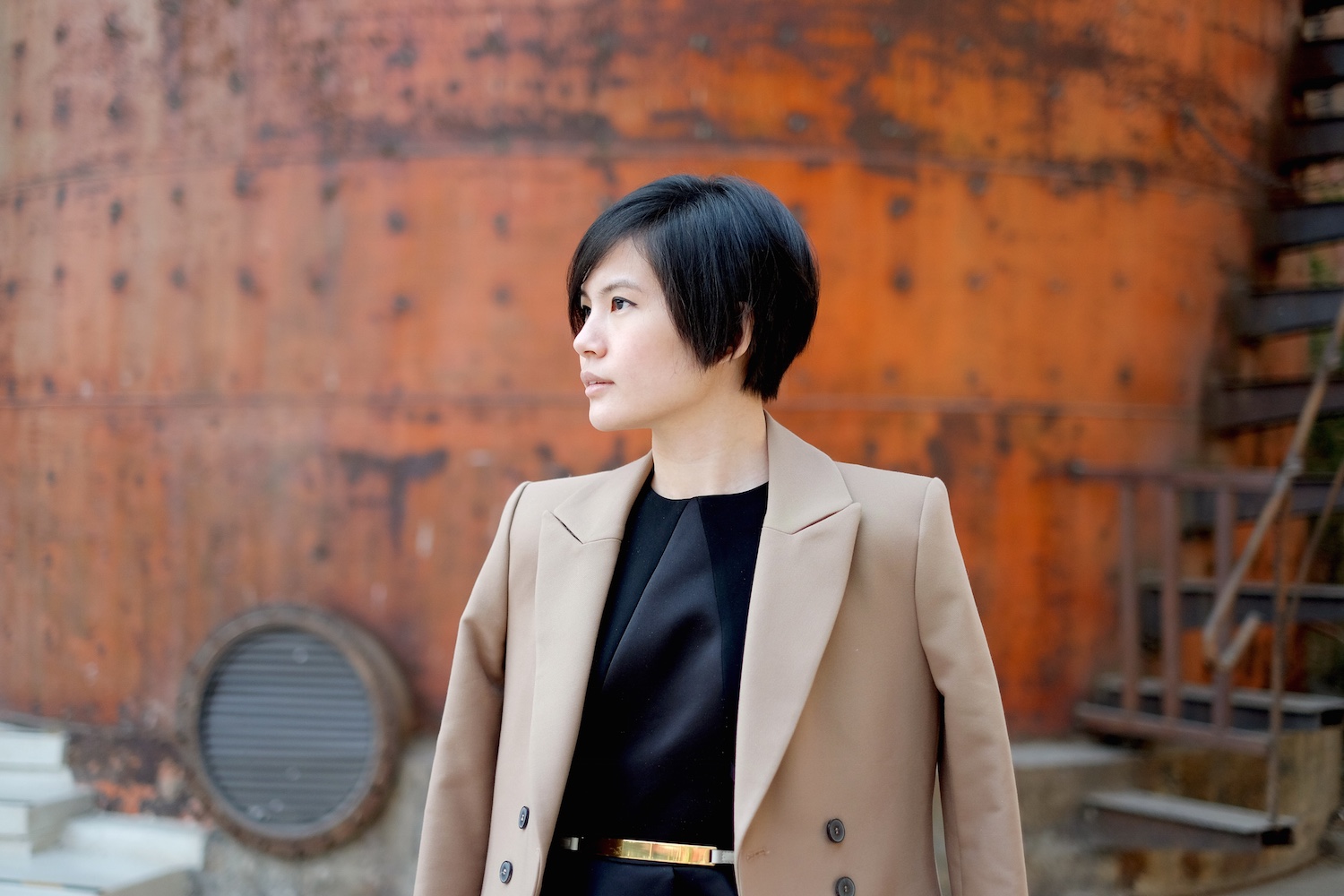 A short haired woman with sepia colored suit and black blouse is posing in front of factory building